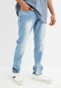 Jeans de mezclilla azul claro con un corte relajado, lavado desgastado, bolsillos laterales y un diseño de pierna recta, combinados con zapatillas blancas y una camiseta gris.