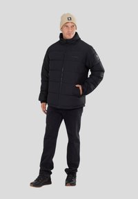 Doudoune noire avec un col montant, des coutures horizontales et une fermeture éclair. Portée avec un jean sombre et des chaussures noires ; un bonnet beige sur la tête.