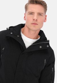 Giacca nera con cappuccio in tessuto testurizzato, con cerniere frontali e dettagli in metallo argentato. Presenta una vestibilità casual e un design con collo alto.