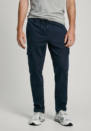 Pantalons cargo navy avec taille à cordon, deux poches latérales et un tissu lisse et léger. Associés à des baskets blanches.