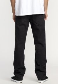 Pantalons en denim noir à coupe décontractée, avec des poches arrière au design arrondi et un léger dégradé. Portés avec des baskets noires.