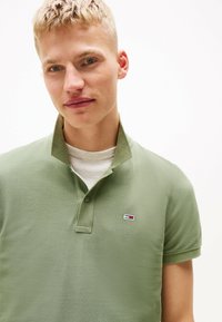 Camisa polo de tejido verde claro con superficie texturizada, con cuello abotonado y pequeño parche de logo en el lado izquierdo del pecho.