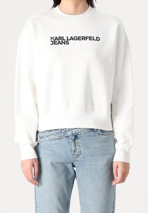 Witte longsleeve trui met zwarte tekst "KARL LAGERFELD JEANS", gedragen met lichtblauwe hoge taille jeans.