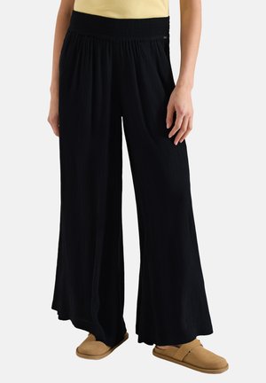 Femme portant un pantalon large noir à taille élastique, des chaussures beige sans lacets, et un haut jaune clair, debout sur un fond blanc.