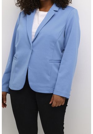 Femme portant un blazer bleu clair sur une chemise blanche et un pantalon noir, debout devant un fond blanc uni.