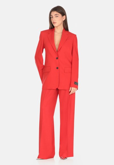 Completo rosso composto da un blazer sartoriale con due bottoni e pantaloni a gamba larga. Realizzato in tessuto liscio con una leggera lucentezza e due tasche.
