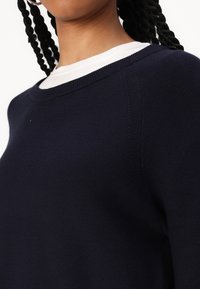 Frau mit geflochtenem Haar, die einen marineblauen Pullover über einem weißen Hemd trägt, der die Strickstruktur und das gerippte Kragen-Detail zeigt.
