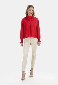 Rote Bluse mit langen Ärmeln und hohem Kragen sowie Knopfdetails, kombiniert mit figurbetonten cremfarbenen Hosen und hautfarbenen High Heels.