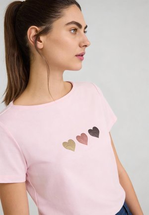 Young woman with brown hair in a ponytail wearing a light pink t-shirt with three metallic hearts in gold, rose, and black on the chest.