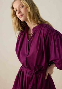 Robe fuchsia avec un col boutonné, des manches volumineuses et une ceinture à nouer à la taille. Fabriquée en tissu léger, elle présente de subtils plis.