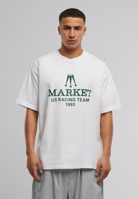 Hvit bomulls t-skjorte med korte ermer, med grønn tekst: "MARKET US RACING TEAM 1993" og et grafisk logo. Enkel, avslappet passform.