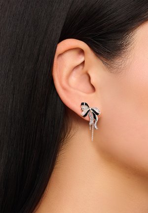Silberne Ohrringe in Form einer Schleife, mit schwarzen Akzenten, klaren Strasssteinen und einem herabhängenden Kettendetail, die nahe am Ohr präsentiert werden.