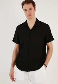 Camisa negra de manga corta con un patrón texturizado en relieve, cuello y cierre frontal con botones. Lleva puesta con pantalones blancos a rayas.