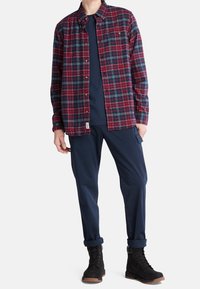Uomo che indossa una camicia a quadri rossi e neri, una t-shirt blu navy, pantaloni cargo blu navy e stivali neri con lacci, in piedi su uno sfondo bianco.
