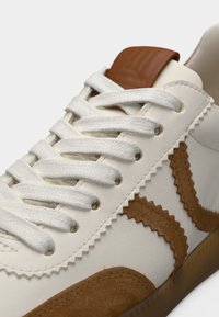 Λευκό αθλητικό παπούτσι με καφέ σουέντ λεπτομέρειες και ραφές σε ζιγκ-ζαγκ. Διαθέτει λευκά κορδόνια και καφέ ταμπελάκι στη φτέρνα με υφή. Ορατή διάφανη σόλα από καουτσούκ.