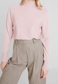 Maglione a coste rosa chiaro con colletto alto, abbinato a pantaloni beige a vita alta con pieghe e vestibilità su misura.