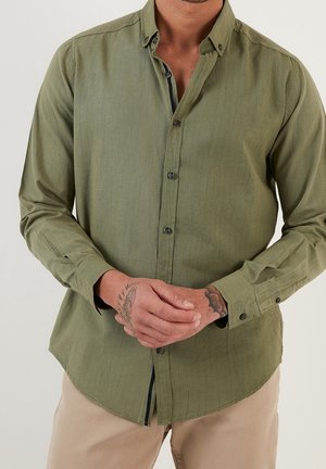 Hombre con una camisa de manga larga color verde oliva abotonada con tatuajes en ambas manos, entrelazadas al frente.