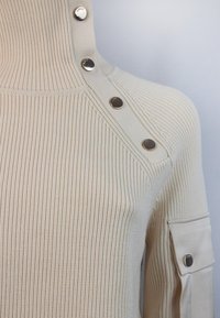 Maglione a coste beige con collo alto, caratterizzato da bottoni a pressione argentati su una spalla e un dettaglio tasca sulla manica.