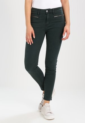 Kobieta ubrana w ciemnozielone, dopasowane spodnie ze kieszeniami na suwak, białe sneakersy i biały top, stojąca ze skrzyżowaną jedną nogą.