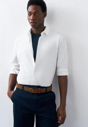 Άνδρας που φοράει λευκό πουκάμισο με κουμπιά και τα μανίκια γυρισμένα, μπλε παντελόνι και καφέ ζώνη, στέκεται μπροστά σε έναν απλό λευκό τοίχο.