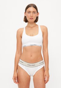 Bijela pamučna grudnjak s niskim izrezom i elastičnom trakom koja nosi crni "Calvin Klein" logo. Uparen s bijelim pamučnim gaćicama i bež akcent trakom.