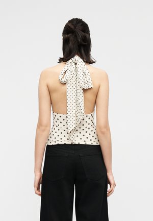 Γυναίκα με σκούρα μαλλιά που φοράει λευκό τοπ με μαύρα πουά, ανοιχτή στην πλάτη και δεμένη στο λαιμό, καθώς και μαύρο παντελόνι, εμφανιζόμενη από πίσω.