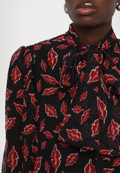 Blouse noire avec une finition texturée, ornée de motifs de feuilles rouges délimités en blanc. Détail de col noué qui ajoute de la dimension au design.