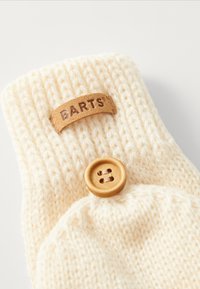 Gestrickte cremefarbene Handschuhe mit einem Lederschild, auf dem "BARTS" steht, und einem großen Holzknopf, der ein strukturiertes Muster aufweist.