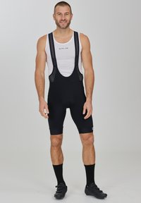Schwarze Radsport-Bib-Shorts mit Netzträgern, einem gepolsterten Sitzpolster und einem eleganten Design. Getragen mit einem weißen ärmellosen Oberteil und schwarzen Radsportschuhen.