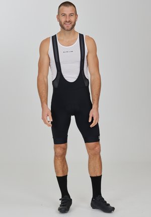 Schwarze Radsport-Bib-Shorts mit Netzträgern, einem gepolsterten Sitzpolster und einem eleganten Design. Getragen mit einem weißen ärmellosen Oberteil und schwarzen Radsportschuhen.