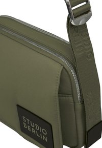 Oliven grøn crossbody taske med lynlås, tekstureret stof og et logo patch. Justerbar skulderrem med mønstret detaljering.