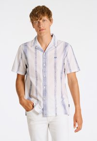 Camisa de manga corta con botones en blanco con rayas verticales azules y patrones en zigzag. Presenta un cuello y bolsillos tipo parche en la parte delantera.
