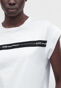 Κοντινό πλάνο ενός ατόμου που φοράει λευκό αμάνικο μπλουζάκι με μαύρη λωρίδα με την επιγραφή "EA7 Emporio Armani" στο στήθος.