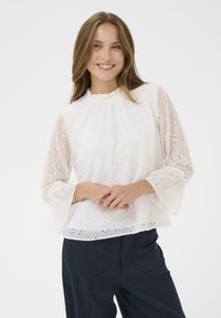 Blouse blanche avec broderie florale, col  à volants et manches évasées. Tissu léger, texture semi-transparente, portée avec un pantalon foncé.