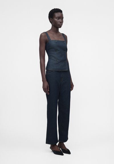 Γυναίκα με αμάνικο σκούρο τζιν τοπ με διπλή σειρά κουμπιών, τζιν παντελόνι με ίσια γραμμή και ψηλή μέση, και μαύρες μυτερές γόβες, όρθια.