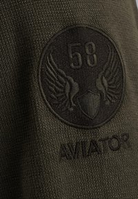 Maslinasto zeleni teksturirani materijal s kružnom crnom izvezenom oznakom s krilima, brojem "58" i riječju "AVIATOR."
