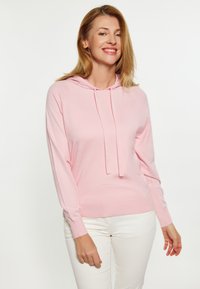 Hanorac roz deschis din tricot texturat, cu detaliu de șnur la gât și mâneci lungi, combinat cu pantaloni albi.