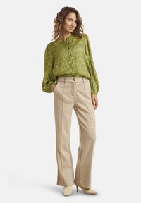 Blusa verde con texture e bottoni sul davanti, abbinata a pantaloni beige a gamba larga con cuciture verticali e décolleté di colore chiaro con punta appuntita.