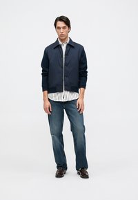 Σκούρο μπλε bomber jacket με κουμπιά, φορεμένο πάνω από ένα ριγέ πουκάμισο. Συνδυασμένο με ξεθωριασμένα μπλε τζιν και σκούρα παπούτσια, στέκεται μπροστά σε λευκό φόντο.