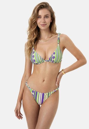 Donna con capelli biondi ondulati che indossa un bikini verde, viola e bianco con motivo, con collana e braccialetto d'oro, in piedi davanti a uno sfondo semplice.