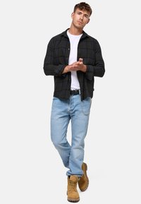 Camisa de franela a cuadros en negro y verde, camiseta blanca debajo, jeans de mezclilla azul claro, botas beige. Incluye un cinturón y un detalle de cadena.