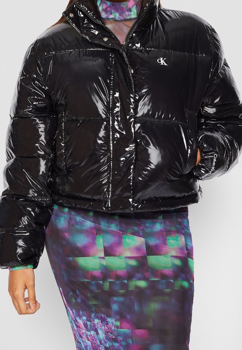 Personne portant une veste matelassée noire brillante avec le logo "cK" et une jupe et un haut à motifs multicolores aux teintes vertes, violettes et bleues.