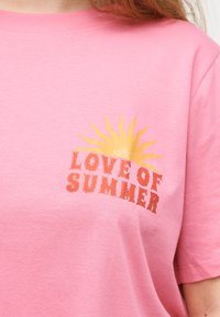 Ροζ μπλουζάκι με γραφικό κίτρινο ήλιο πάνω από το κείμενο "ΑΓΑΠΗ ΤΟΥ ΚΑΛΟΚΑΙΡΙΟΥ" στο στήθος, φορεμένο από ένα άτομο με κόκκινα μαλλιά.