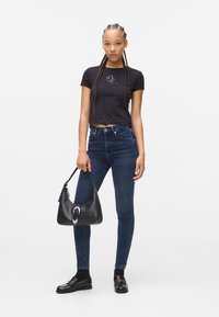 Tricou scurt negru cu logo, blugi skinny de culoare bleumarin, geantă de umăr din piele neagră cu accent metalic și mocasini negri cu studuri.