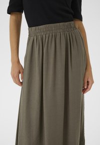 Jupe longue vert olive avec une taille élastique et un tissu doux et texturé. Présente des plis pour le mouvement. La partie supérieure n'est pas visible sur l'image.