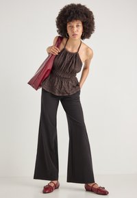 Haut halter plissé marron avec taille froncée, associé à un pantalon large noir et des chaussures plates bordeaux. Sac fourre-tout rouge porté sur l'épaule.