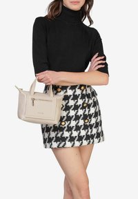Beige struktureret håndtaske med to top håndtag, lynlåslomme foran og diskret branding. Modellen har en sort top og et houndstooth-skirt på.