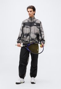 Νεαρός άνδρας που φοράει μαύρη και γκρι ριγέ φόρμα, κρατώντας ρακέτα του τένις και μπάλα, στέκεται μπροστά σε απλό φωτεινό φόντο.