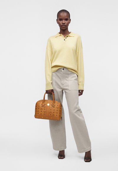 Femme debout face à l'avant, portant un pull jaune pâle, un pantalon beige clair, tenant un sac à main marron à motifs et des sandales noires à bout ouvert.