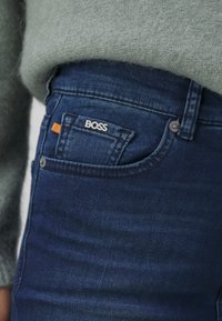 Jeans de denim azul oscuro con corte ajustado, que presentan bolsillos frontales y un logo plateado "BOSS" en el bolsillo trasero, complementados con una etiqueta naranja.